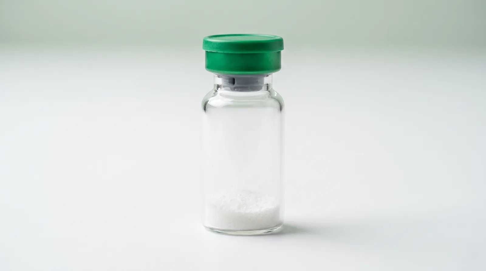 Einzelnes Vial mit grünem Verschluss und weißem lyophilisiertem Pulver am Boden vor weißem Hintergrund